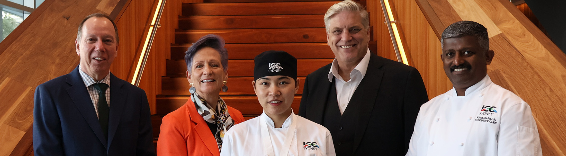 Thida Htay wins culinary gold, pictured holding her certificate with ICC Sydney leaders standing next to her against dark timber stairs.
