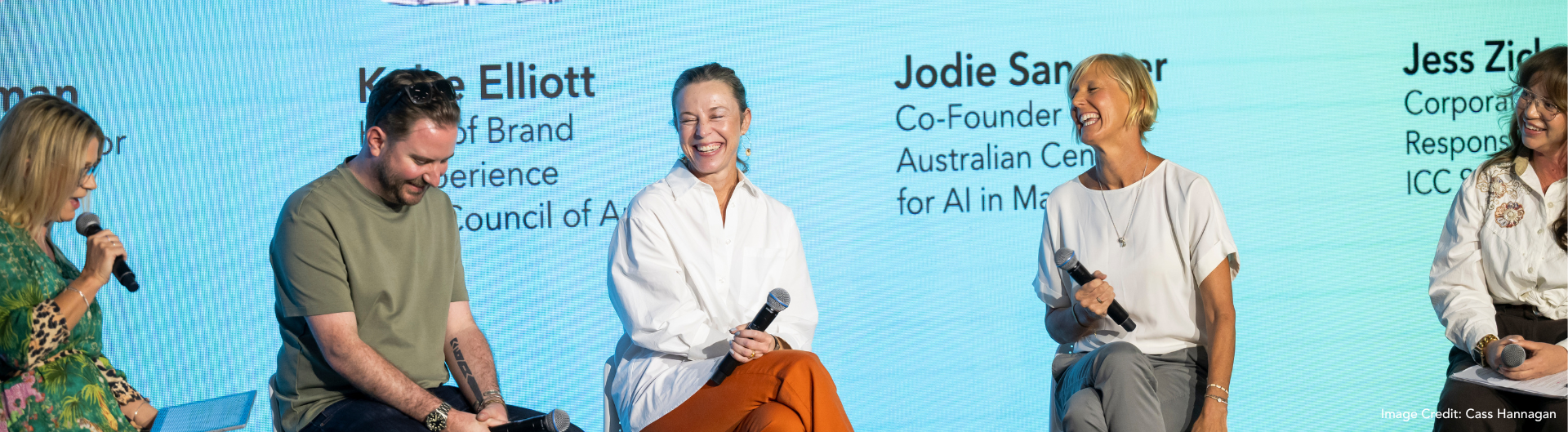 five panel members sit on stools in front of a blue screen laughing with each other