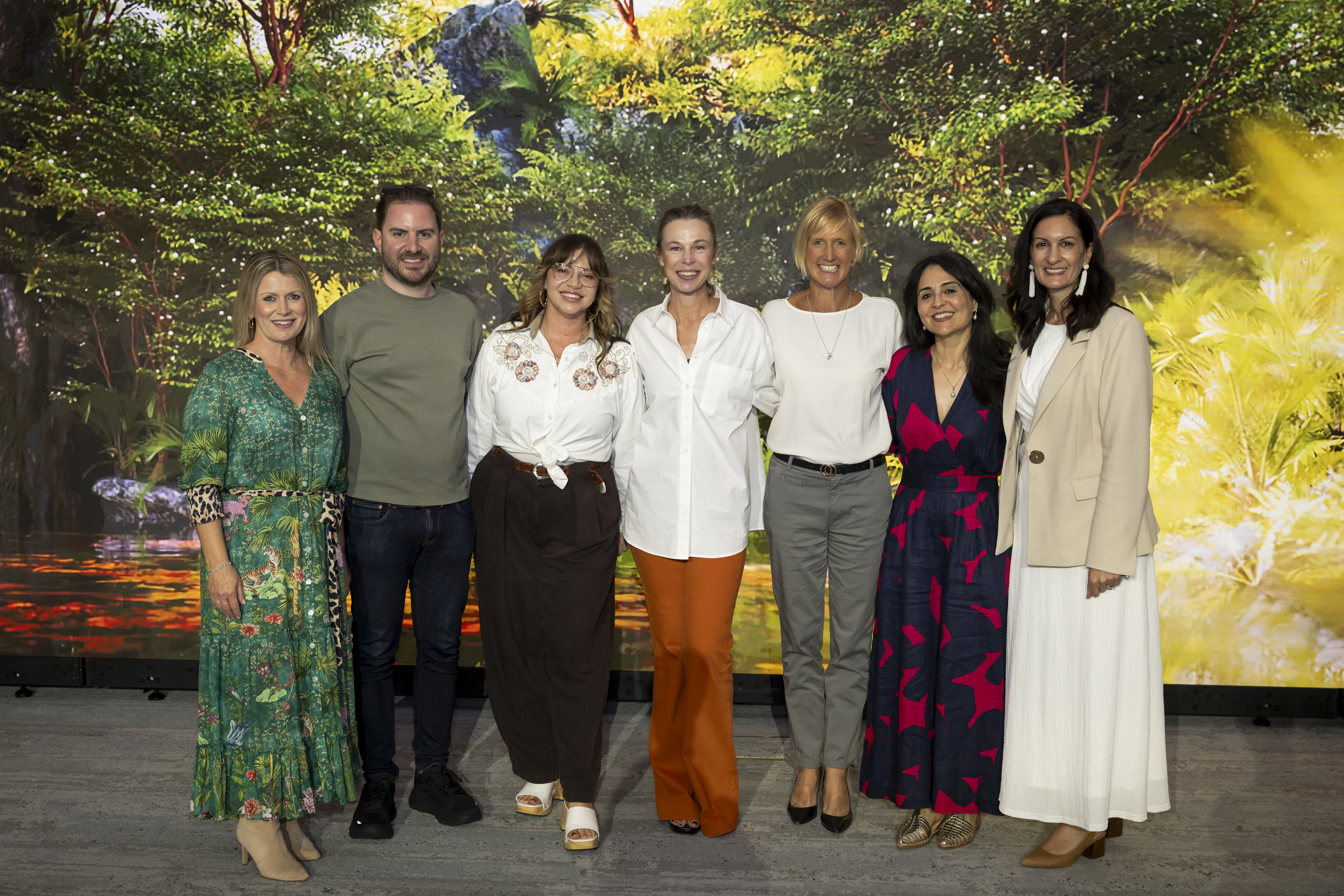 six people standing in front of an image of leafy green trees and smiling at the camera